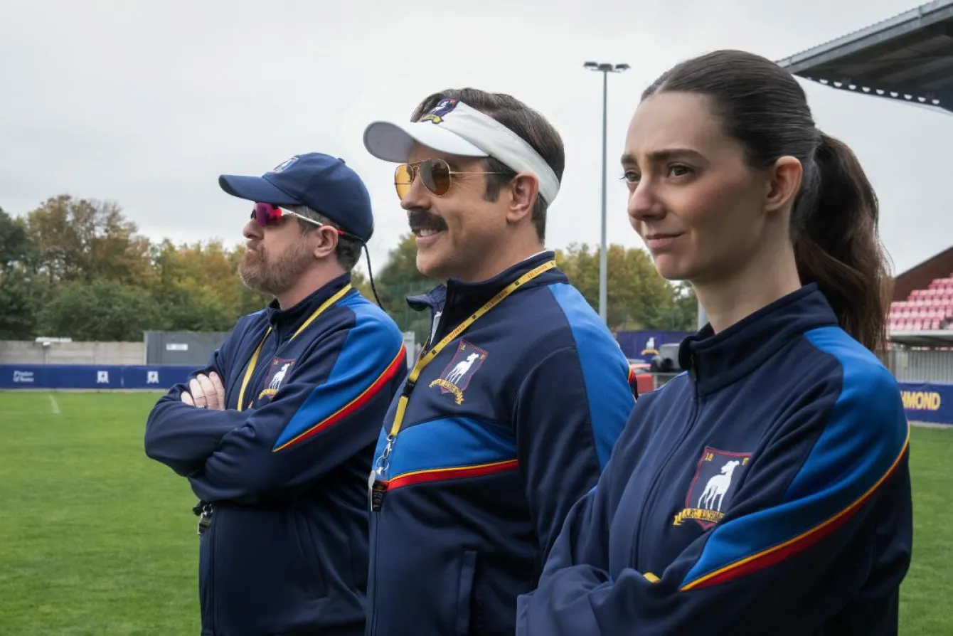 Brendan Hunt como Coach Beard, Jason Sudeikis como Ted Lasso e Tanya Reynolds como Alice Chilton em cena da 4ª temporada de Ted Lasso. Na nova fase da série, Ted retorna ao AFC Richmond para comandar a equipe feminina do clube.