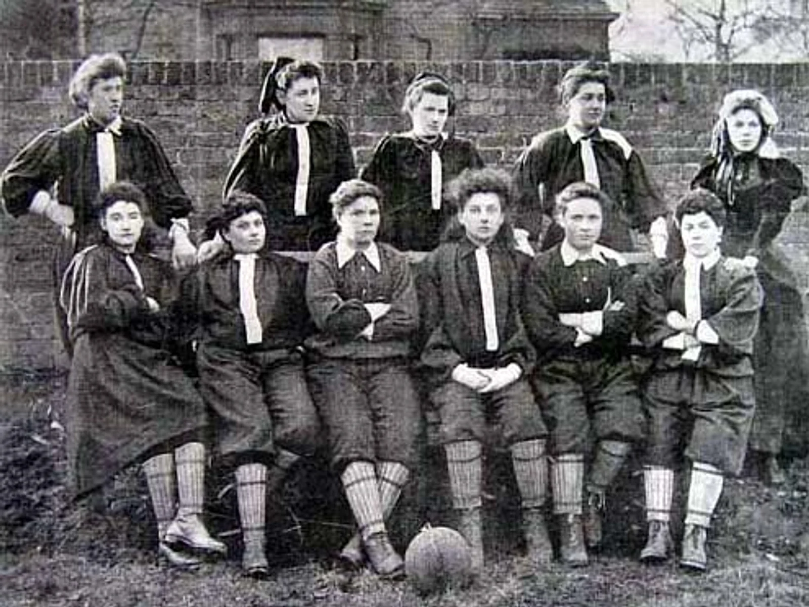 Fotografia de 1895 com as onze jogadoras do time do Norte do British Ladies' Football Club posadas diante de um muro de tijolos, de blusa escura, calção e meiões, com uma bola de couro no chão