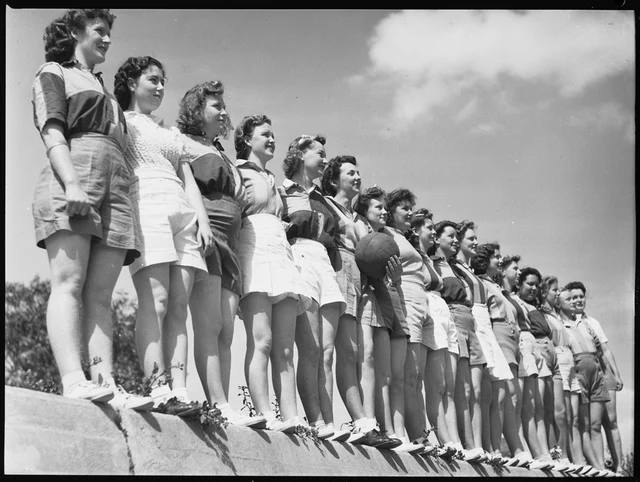 Fotografia de 1942 com cerca de dezoito jogadoras do time feminino de Granville perfiladas sobre um muro baixo contra o céu, uma delas segurando uma bola de couro