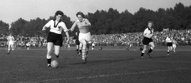 Fotografia em preto e branco de 1956: uma jogadora alemã conduz a bola perseguida por uma holandesa, com a arquibancada lotada ao fundo