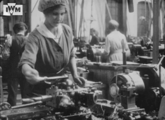 Fotograma de 1917: uma operária de touca opera um torno numa fábrica de munição da Vickers, com outras mulheres trabalhando ao fundo