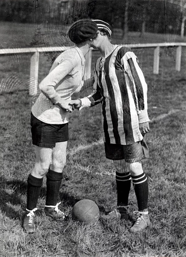 Fotografia de 1920 com duas capitãs de times femininos se cumprimentando no gramado antes do jogo, uma de camisa lisa e outra de camisa listrada, com a bola de couro no chão entre elas