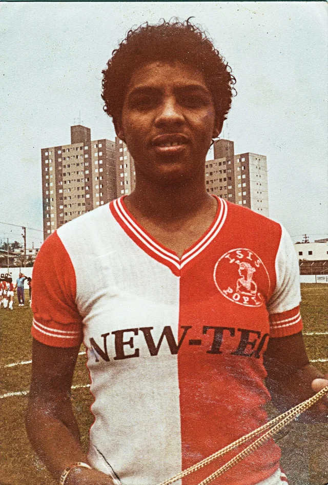 Retrato colorido de 1984: Marileia dos Santos, de frente, com a camisa vermelha e branca do Isis Pop, num campo em São Paulo com prédios ao fundo